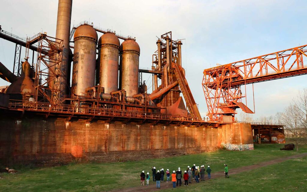 Rust-hued steelwork equipment dwarfs a cluster of people wearing hardhats in the foreground.